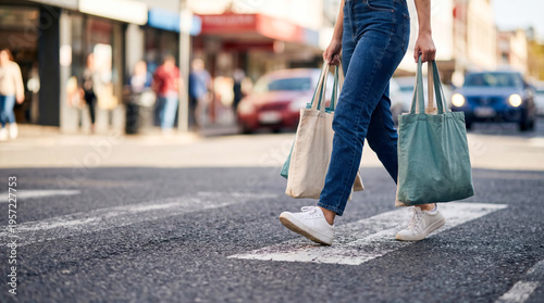 A woman walks across a crosswalk holding two shopping bags. Cars are parked nearby and people are seen walking along the street. It is a busy afternoon in the shopping area