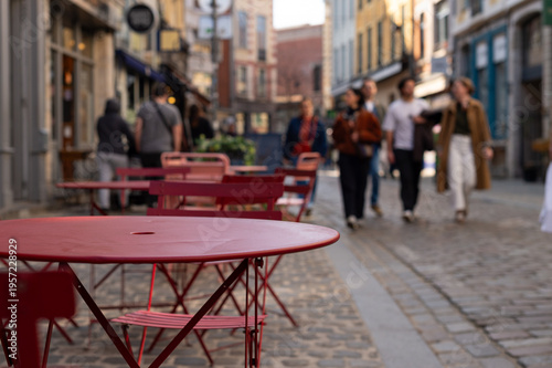 Canvastavla Red metal tables and chairs at an outdoor cafe on a cobblestone street in Lille, France, with blurred people walking in the background
