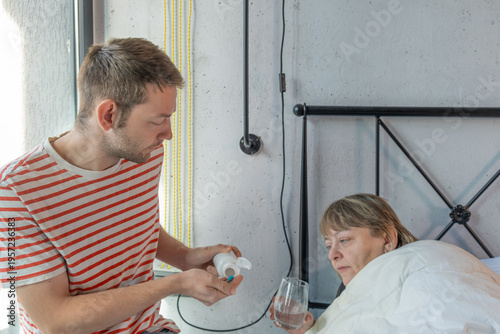 Adult son helps his sick mother in bed by giving medicine and water at home. Family care for elderly parents, support and responsibility during illness and recovery. Caregiving and home healthcare