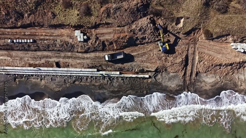 Workers are seen operating machinery and laying materials along the shoreline. Waves crash on the beach as construction progresses on a building project.