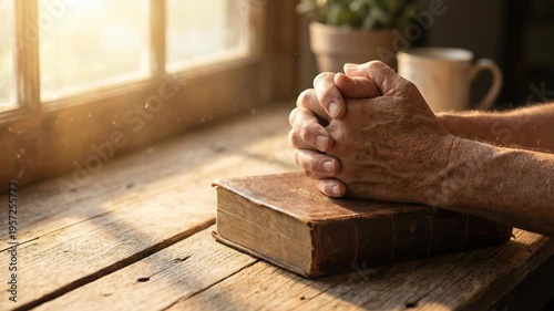 Senior man praying with folded hands on holy bible. Devotional meditation at wooden table near window. Spiritual practice and faith in god, christianity concept.