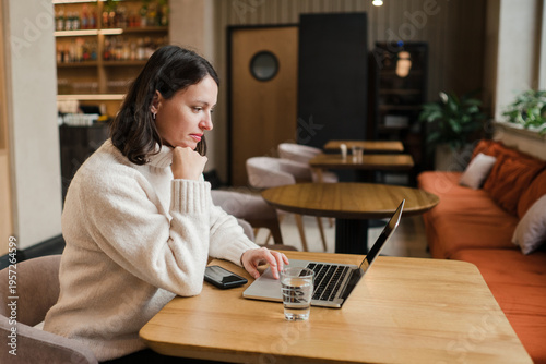Caucasian woman in her 40s with dark hair wearing white knit sweater working in coworking space using her laptop. Mature female spending time in modern office completing creative or routine tasks.
