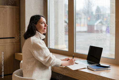 Caucasian woman in her 40s with dark hair wearing white knit sweater working in coworking space using her laptop. Mature female spending time in modern office completing creative or routine tasks.