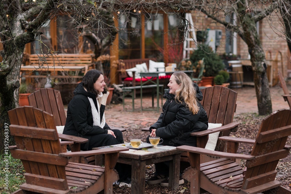 Obraz premium Two pretty caucasian women in their 40s wearing warm clothes spending time outdoors in autumn park cafe. Mature females enjoying personal time, talking, drinking tea, relaxing and breathing fresh air.
