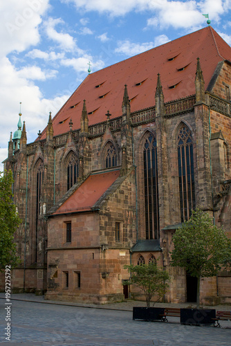 Gothic church rises in Nurnberg under a bright sky. Germany. Tall windows and stone details show long history. A quiet square and trees frame the building in soft light.