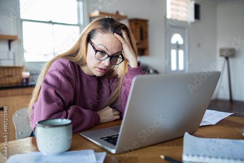 Stressed woman reviewing bills on laptop at home kitchen table