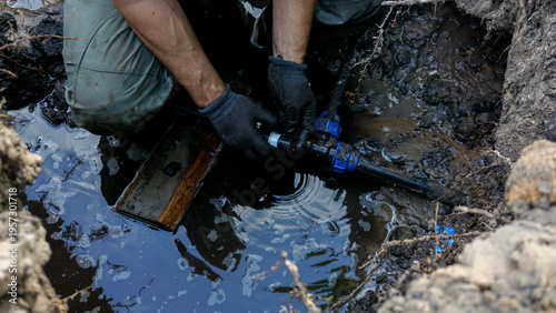 Gloved plumber crouching in a flooded muddy trench replacing a blue compression pipe, fitting by hand during an underground water leak repair on residential property.