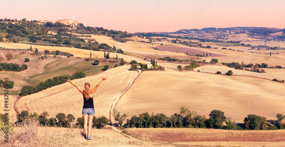 Fototapeta premium Young woman with open arms enjoying the golden sunset over the rolling hills of Tuscany, Italy. Breathtaking landscape captures the iconic rolling hills of Val d'Orcia during the golden sunset