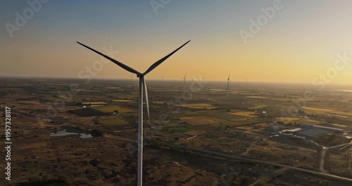 Golden hour aerial view of wind turbines across rural farmland, with rotating blades under a warm orange sky, showcasing clean energy and a serene countryside landscape of Gujrat, India