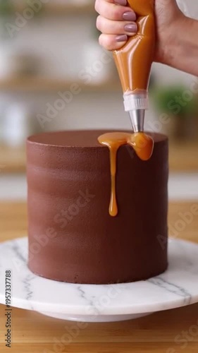 A hand drizzles caramel sauce onto a chocolate cake on a marble plate in a kitchen