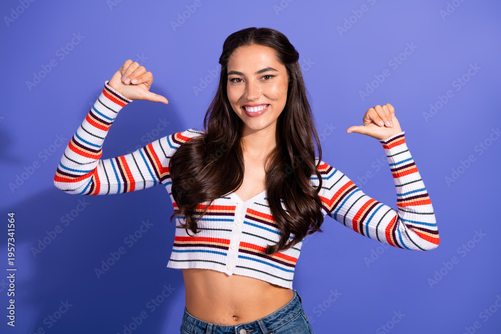 Fototapeta premium Young female model wearing a striped crop top poses with arms raised against a violet background for fashion lifestyle and advertising