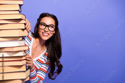 Fotografie Young brunette woman with glasses holding a tall stack of books smiling against