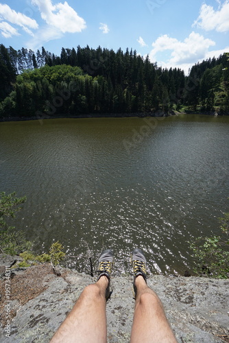 Point of view with legs on rocky edge above water, forest landscape across lake in background