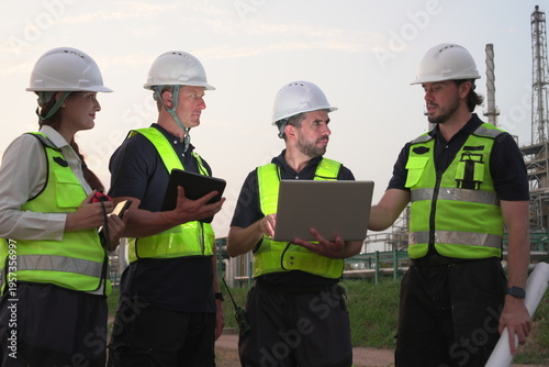A group of male staff, male inspectors, female architects, male foremen, and managers, all wearing uniforms and helmets for safety, were reviewing blueprints, discussing plans at an oil refinery 