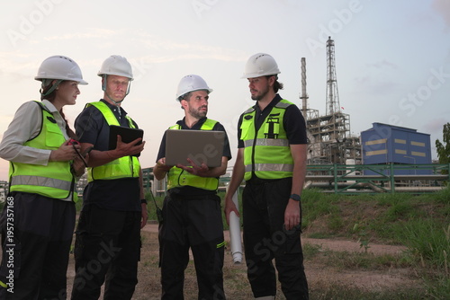 A group of male staff, male inspectors, female architects, male foremen, and managers, all wearing uniforms and helmets for safety, were reviewing blueprints, discussing plans at an oil refinery 