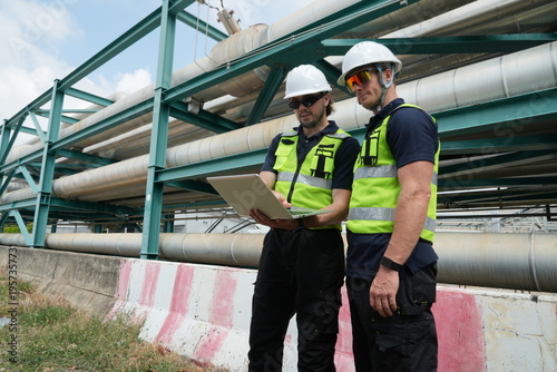 A team of male staff, including male inspectors and foremen in uniforms and helmets, along with engineers reviewing blueprints and planning, were inspecting maintenance work at an oil and petrochemica