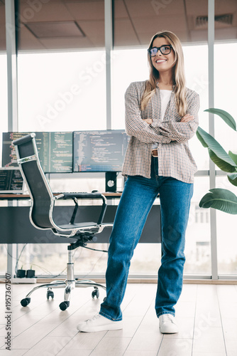 Confident female programmer standing in a modern office workspace with crossed arms in front of computer screens displaying code, casual tech startup developer portrait in bright open-plan studio