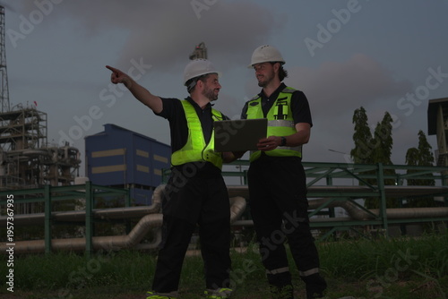 Male engineering staff and a male foreman/manager, all in uniform and helmets, are discussing and planning inspections of maintenance work at an oil refinery and petrochemical plant at night.
