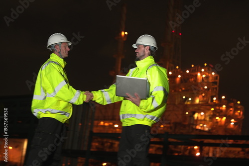 A team of male staff and inspectors shakes hands and congratulates a male foreman in uniform and helmet, an engineer, as the team discusses and plans a nighttime inspection of maintenance work at an o