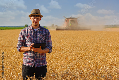 Young farmer agronomist wearing a straw hat controls the wheat harvest on a digital tablet, with combine harvester reaping wheat in the background - close up. Sunny summer day
