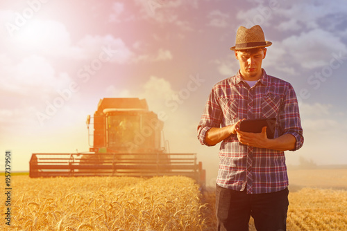 Young farmer agronomist wearing a straw hat controls the wheat harvest on a digital tablet at sunset, with combine harvester reaping wheat. Beautiful rural summer landscape. Food production