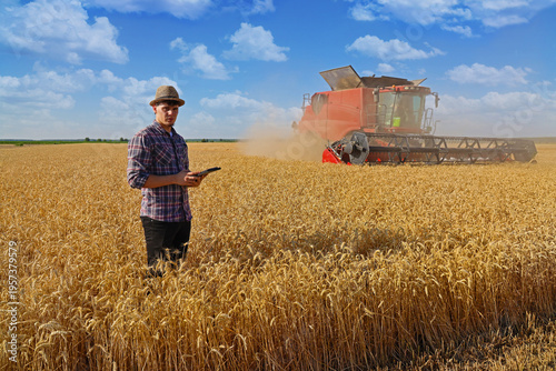 Young farmer agronomist wearing a straw hat controls the wheat harvest on a digital tablet, with combine harvester reaping wheat in the background - close up. Sunny summer day