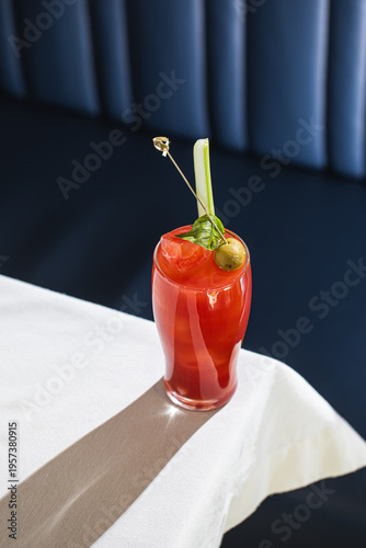 Red bloody mary cocktail with celery olive and ice served in tall glass on table in bar interior