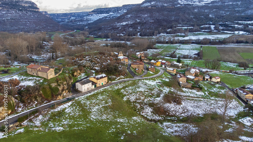 Картината върху платно Aerial view of the small village of Arroyuelos, featuring green meadows dusted with light snow and surrounded by snow-capped mountains