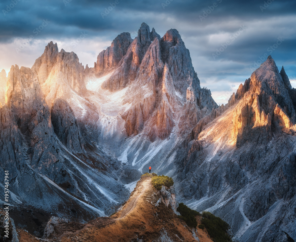 Fototapeta premium Girl on the mountain peak and high rocks at colorful sunset in autumn. Tre Cime, Dolomites, Italy. Colorful landscape with woman on trail, cliffs, grass, cloudy sky in fall. Hiking. Aerial view