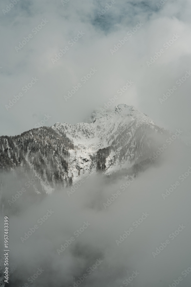 Obraz premium Dense white fog drifts through a rugged alpine valley, partially veiling the massive, snow-covered mountain faces under a heavy, overcast sky.