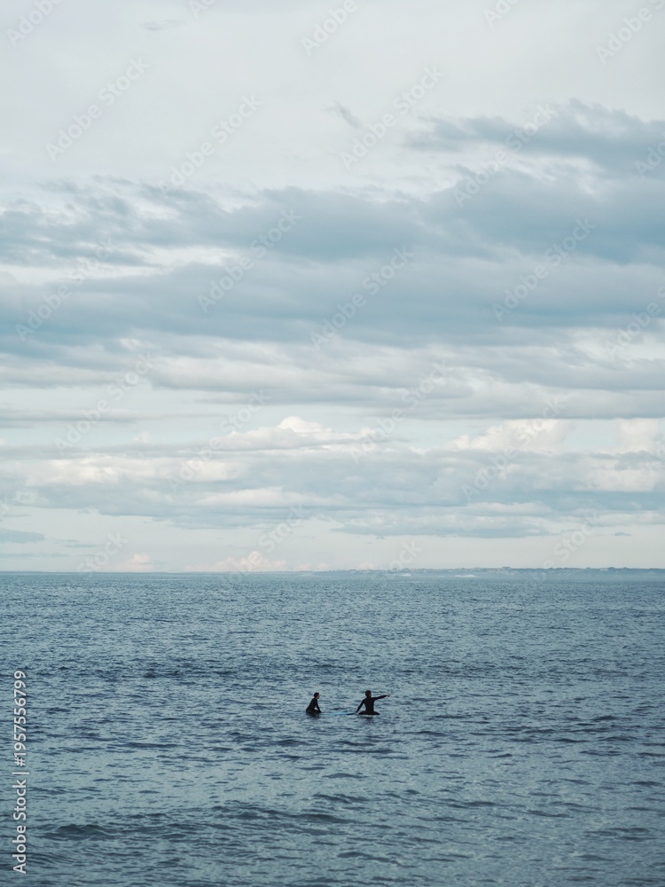 Obraz premium Two surfers are alone at sea, waiting for waves and pointing to the horizon, where perhaps something interesting will happen; meanwhile, the horizon of the sea, the sky and the clouds are calm in the 
