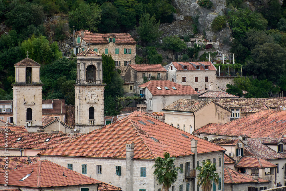 Fototapeta premium View of historic stone buildings and orange tile roofs in the old town of Kotor with a clock tower and church bell tower against a green hillside.