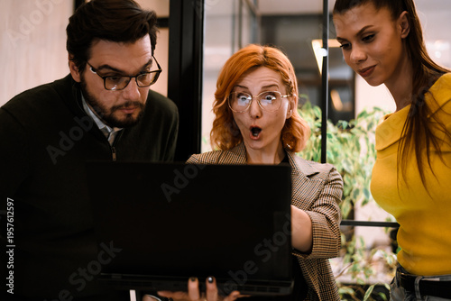 Schilderij op canvas Three colleagues gather around a laptop in a modern office, expressing surprise