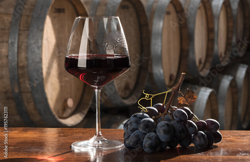 A glass of red wine and fresh grapes rest on a wooden table in a wine cellar with barrels
