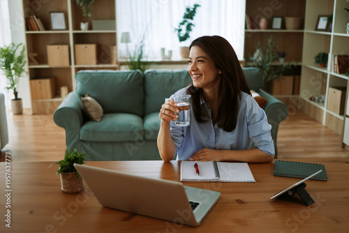 Obraz na plátne Young woman smiling while working from home holding a glass of water at her desk