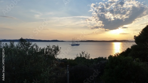 Sailing ship anchored on calm sea at sunset with golden reflections on the water and soft clouds creating a peaceful Mediterranean evening atmosphere