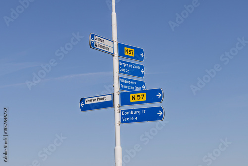 Middelburg, Zeeland, Netherlands. 18 March 2026. Blue Directional Road Signs on White Pole in Zeeland, Pointing to Domburg, Veere, Vlissingen and Goes under Clear Blue Sky.
