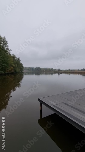 Panorama of a lake in autumn woodland. Pier and green forest reflecting on the calm water. Serene landscape with overcast sky.
