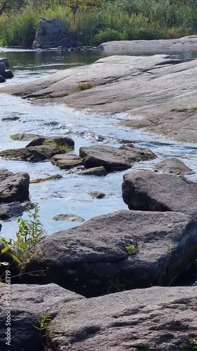 Shallow rocky stream over granite rocks. Vertical video of sunlit stream flowing in granite canyon, clear water ripples over and around dark rocks. Ros river in the Kyiv region.