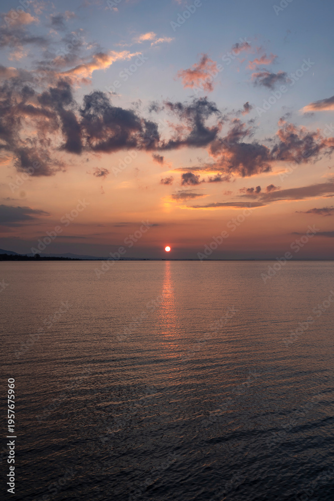 Obraz premium Breathtaking sunset over Lake Constance viewed from the Bregenz shore, featuring a vibrant orange sun dipping towards the horizon with dramatic clouds reflecting in the calm water.