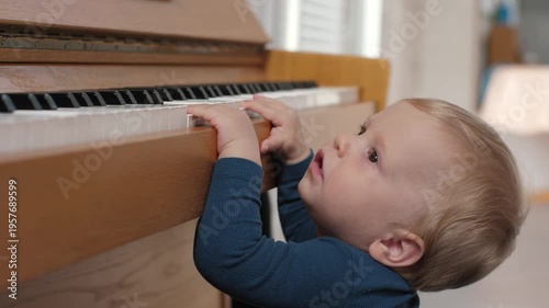 White toddler at piano exploring keys with tiny fingers, showing curiosity and early musical discovery in cozy living room. Little kid fingers press the piano keys, natural light