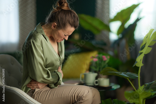 A woman wearing an olive green silk shirt sits in a chair clutching her lower abdomen, depicting stomach pain or digestive distress in a modern living space.