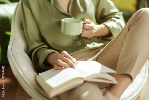 A peaceful scene of a woman in a sage green shirt enjoying a quiet moment of mindful reading and tea in her modern, sun-drenched living room.