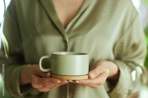 A close-up of a woman in a sage green silk blouse holding a ceramic mug, embodying a minimalist and mindful approach to self-care and morning relaxation at home.