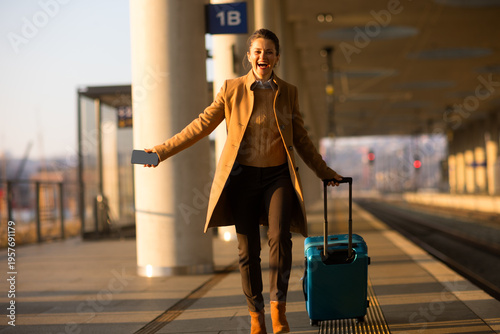 Foto A smiling woman in a tan overcoat walks along a sunlit train platform, carrying a smartphone and pulling her blue rolling luggage during a morning commute
