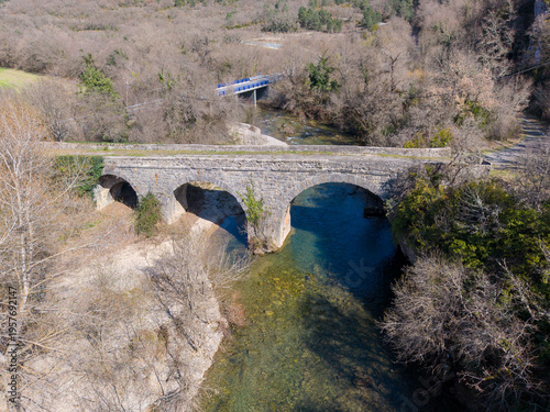 An ancient Roman stone bridge with three arches spans a clear river. The surroundings are natural, with mostly dry or winter vegetation, leafless trees, and a few green shrubs. 