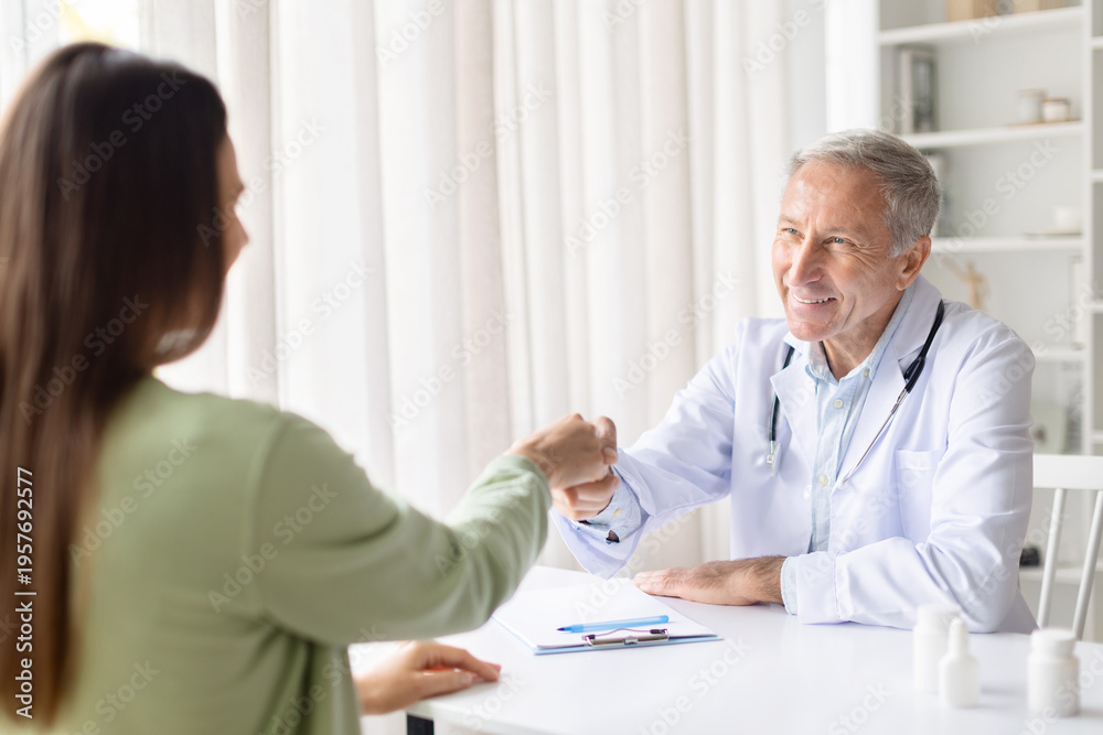 Obraz premium Senior male doctor greeting female patient with friendly fist bump during consultation. Medical professional building trust and positive relationship in healthcare environment