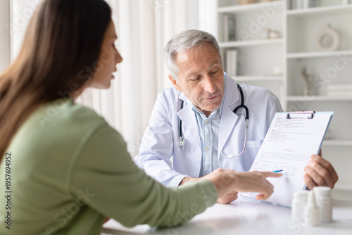 Senior male doctor explaining medical document and treatment plan to female patient during clinic consultation. Healthcare professional reviewing healthcare paperwork