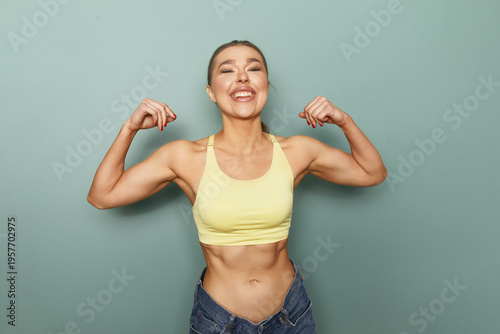 Fit woman shows her muscles in front of a green wall during a workout session at a gym while smiling and expressing confidence in her strength and fitness journey