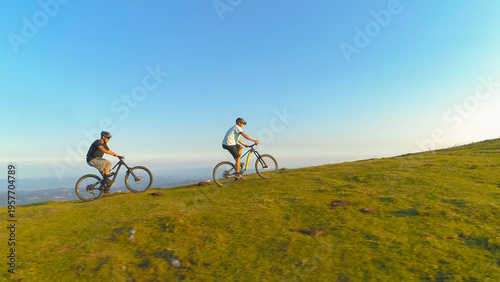 Obraz na plátně DRONE: Two mountain bikers ride their electric bicycles up a steep grassy hill